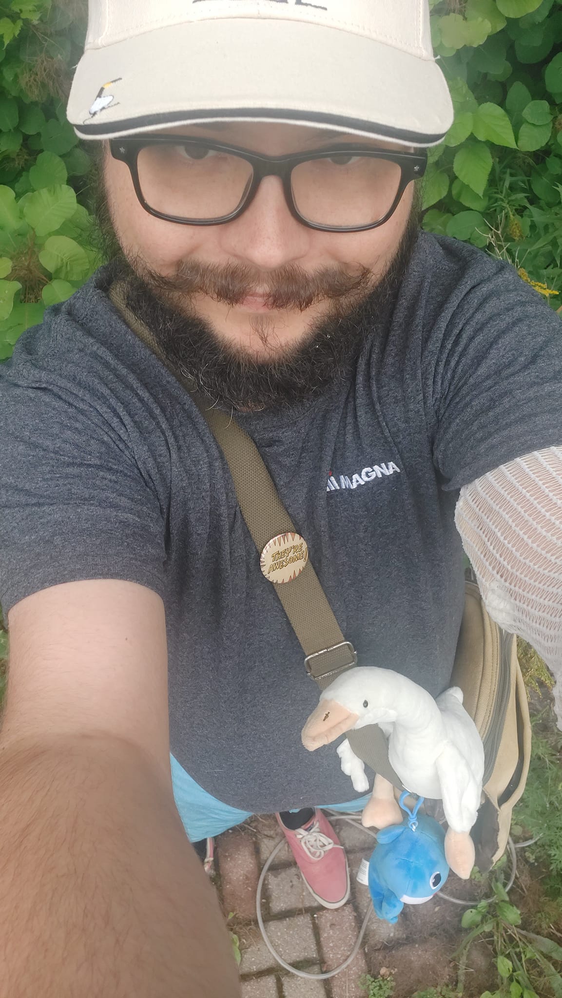 A man with short hair and bushy facial hair stands, the camera is angled so you can see his messanger bad with a blue shark and white goose plush attached